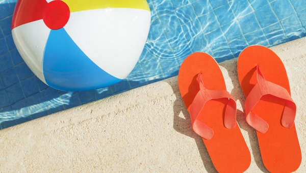 Zwei orange Flipflap-Sandalen am Poolrand, im Wasser schwimmt ein farbiger Ball